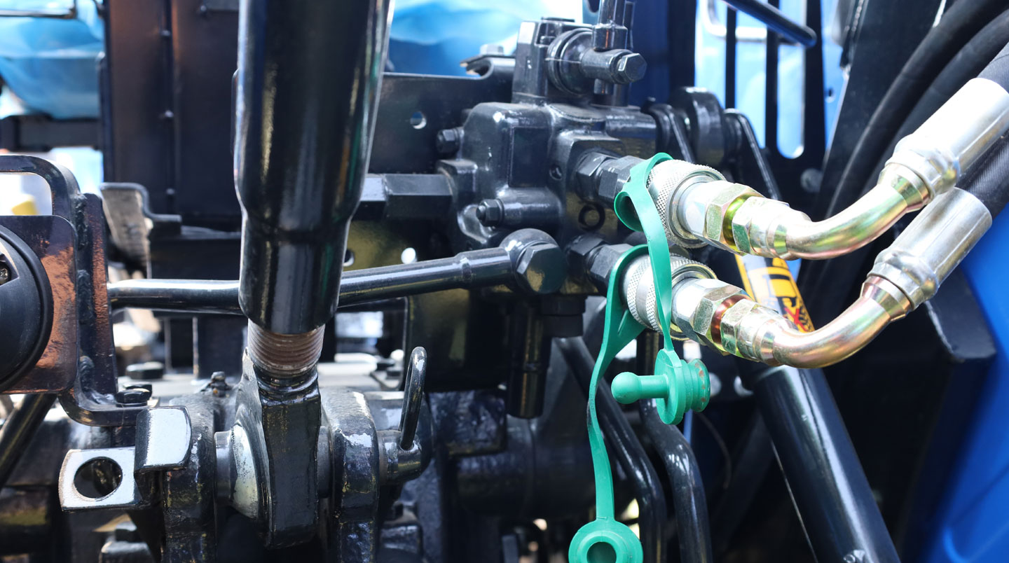 Gros plan distributeurs hydrauliques et flexibles haute pression sous châssis tracteur agricole, révélant traces potentielles fuites huile sur raccords métalliques